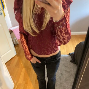 Vinröd spetsblus med volangdetaljer - Vinröd blus i transparent spets med långa ärmar och croppad passform. Blusen har volangdetaljer vid axlarna och fint spetsmönster. Den har en knapp i nacken som gått sönder men det är inget som märks. 🩷