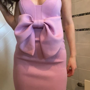 Ljus rosa/lila kort klänning med rosett från ASOS - Supersöt ljusrosa klänning från ASOS med stor rosett framtill. Klänningen är kort och har en figurnära passform med axelbandslös design. Materialet är mjukt och känns lyxigt mot huden, perfekt för att sticka ut med en unik look.