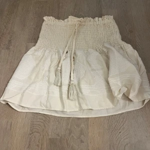 Beige kortkjol med smock från Zara - Supersöt beige kortkjol från Zara med smockad midja och knytband med tofsar. Kjolen har volangig nederdel och ett diskret rutigt mönster i strukturen. 