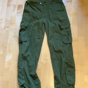 Gröna cargopants med snörning, M - Säljer ett par gröna cargopants i storlek M med flera stora fickor på benen och bak. Byxorna har snörning i midjan och är gjorda av bomull, viskos och spandex för skön passform. Perfekta för dig som gillar streetstyle och funktionella detaljer.