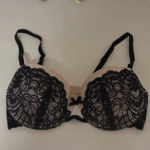 Svart spets bh från Hunkemöller - Supersnygg bh från Hunkemöller i storlek 75B. Nude bas med svart spets och blommigt mönster, samt en liten rosett framtill. Axelbanden är justerbara och ryggen har klassisk hakknäppning. Perfekt för dig som gillar detaljer och vill ha något extra.