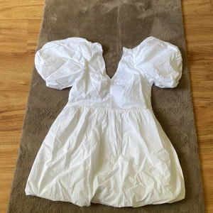 Vit puffärm klänning från Gina Tricot - Supersöt vit kortklänning med puffiga kortärmar och v-ringad hals. Baktill finns en snygg knyt-detalj som ger extra stilpoäng. Klänningen är gjord i bomull och har en luftig, voluminös kjol som ger en cool och trendig look.