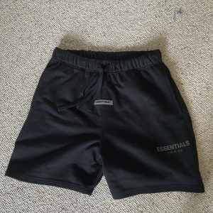 Svarta shorts från Fear of God Essentials - Svarta shorts från Fear of God Essentials med elastisk midja och snörning. Klassisk och clean design med logga framtill. Perfekta för chill dagar eller när du vill ha en avslappnad look. Mjukt material som känns skönt mot huden.