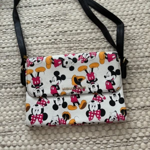 Michael Kors axelväska Disney print - Snygg axelväska från Michael Kors med färgglatt Disney-motiv av Musse och Mimmi Pigg. Väskan är vit med svarta, rosa och gula detaljer, har svart axelrem och flera fack med dragkedja. Perfekt för dig som vill sticka ut med en lekfull och trendig accessoar. Köpt till spex så använd 1-2 gånger. Köpt för 500kr. Pris kan diskuteras.