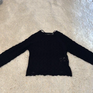 Svart stickad tröja från Vero Moda - Svart stickad tröja från Vero Moda med långärmad design och snyggt hålmönster. Tröjan har rund halsringning och vågig kant nertill och vid ärmslut. Liten i storleken. Aldrig använd