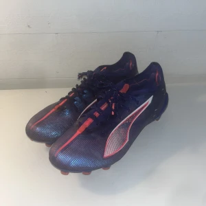 Puma blå fotbollsskor med neonrosa detaljer - Säljer ett par snygga fotbollsskor från Puma i mörkblått med neonrosa och vita detaljer. Skorna har snörning, låg profil och mönstrad ovandel i syntetmaterial. Perfekta för dig som vill sticka ut på planen med stilrena och sportiga skor.