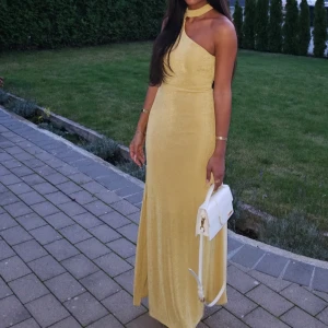 Gul oneshoulder långklänning - Säljer en elegant gul långklänning med oneshoulder-design och öppen rygg. Klänningen har en smickrande tight passform och ett mjukt, glansigt material som ger en lyxig känsla. Perfekt för bal eller fest, med snygg detalj runt halsen.