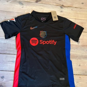 FC Barcelona Lewandowski Nike tröja - Helt ny FC Barcelona matchtröja med Lewandowski och nummer 9 på ryggen. Svart bas med röda och blå sidopaneler, röd Nike-logga och Spotify-tryck på bröstet. Perfekt för dig som älskar Barca.