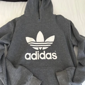 Grå Adidas hoodie med logga - Säljer en grå hoodie från Adidas med stor vit trefoil-logga och Adidas-tryck på bröstet. Tröjan har huva, ribbade muddar och är långärmad. Materialet är mjuk bomullsmix som är perfekt för chill eller träning. Klassisk streetwear-stil som alltid funkar.