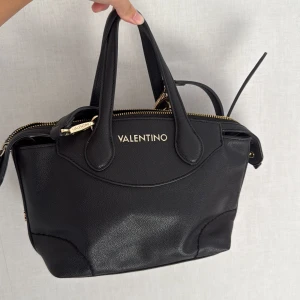 Svart handväska från Valentino - Snygg svart handväska från Valentino med guldfärgade detaljer och dragkedja. Väskan har både handtag och justerbar axelrem.  Tillverkad i skinn. 