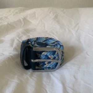 Blått flätat bälte från ONEWear - Snyggt flätat bälte från ONEWear Sweden i olika blå nyanser, beige och vitt. Bältet har detaljer i mörkblått skinn och ett silvrigt spänne. Perfekt accessoar för att lyfta din outfit med lite färg och stil.