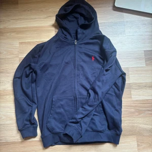 Mörkblå hoodie från Polo Ralph Lauren - Mörkblå hoodie från Polo Ralph Lauren med dragkedja och huva. Klassisk röd logga broderad på bröstet och känguruficka framtill. Tillverkad i mjuk bomull och har ribbade muddar vid ärmslut och nederkant. Custom fit för en snygg passform.