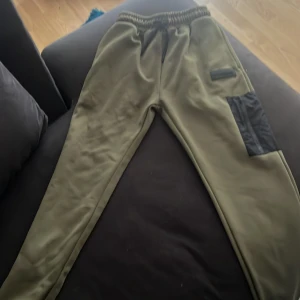 Olivgröna cargopants från McKenzie S - Olivgröna cargopants från McKenzie med svart patchficka på benet och elastisk midja med snörning. Byxorna har en loose fit och är tillverkade i mjukt material, perfekta för chill dagar. Snygg streetwear-vibe med diskret logga på benet.