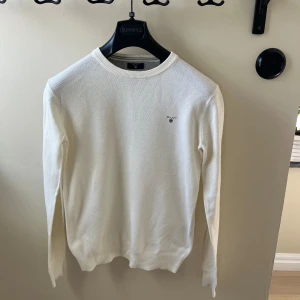Vit stickad tröja från GANT - Vit stickad tröja från GANT med diskret logga på bröstet. Tröjan har rund halsringning, ribbade muddar och lång ärm. Perfekt för dig som gillar en clean och klassisk stil. Materialet är mjukt och bekvämt.
