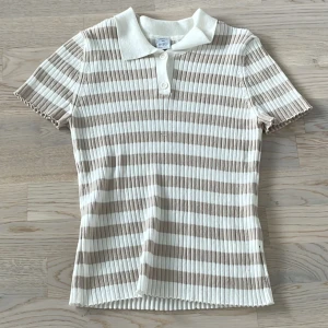 Beige och vit randig pikétröja - Superfin ribbad pikétröja från Lindex, knappt använd i beige och vitt med klassisk krage och knappar framtill. Tröjan har korta ärmar och är figurnära i modellen. Perfekt för en fräsch och stilren look i vår och sommar.