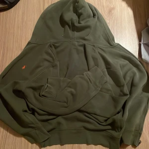 Olivgrön hoodie från Polo Ralph Lauren - Snygg olivgrön hoodie från Polo Ralph Lauren med klassisk huva och dragkedja. Tröjan har en liten orange broderad logga på bröstet och är tillverkad i mjuk bomull. Perfekt för en chill och avslappnad stil.