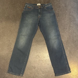Wrangler blå raka jeans - Klassiska blå jeans från Wrangler med raka ben och snyggt slitna detaljer. Jeansen har fem fickor, bruna sömmar och läderpatch på bakfickan. Perfekt för dig som gillar en tidlös och avslappnad look.