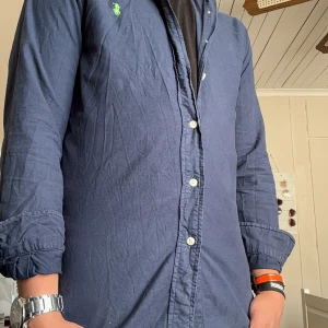 Blå skjorta Polo Ralph Lauren slim fit - Snygg marinblå skjorta från Polo Ralph Lauren i slim fit-modell. Skjortan har långa ärmar, klassisk krage och knäppning framtill. Broderad grön logga på bröstet ger en stilren touch. Perfekt för dig som gillar en clean och tidlös look. Skick 9/10.    Det är bara höra av sig om de är något du undrar över 😇priset är inte hugget i sten🤩 