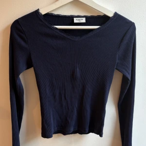 Mörkblå ribbad topp från Kappahl - Snygg mörkblå långärmad topp från Kappahl med ribbad struktur och v-ringning. Fina spetsdetaljer längs halsringningen ger en extra touch. Perfekt att styla med jeans eller kjol för en trendig look.