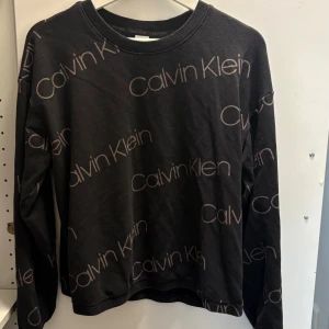 Svart Calvin Klein sweatshirt M - Svart sweatshirt från Calvin Klein med stort logomönster över hela tröjan. Långärmad och i mjuk bomull, perfekt för en chill och stilren look. Rund halsringning och relaxed passform.