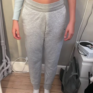 Grå mjukisbyxor från Puma - Snygga ljusgrå mjukisbyxor från Puma med svart logga på benet. Byxorna har bred resår i midjan, ribbade muddar vid bensluten och praktiska sidofickor. Perfekta för chill eller träning, tillverkade i mjuk bomullsblandning för maxad komfort.