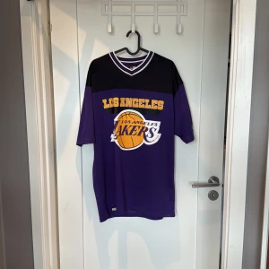 Los Angeles Lakers tröja mesh - Säljer en sportig Los Angeles Lakers tröja i meshmaterial med klassisk lila och svart färg, stora gula tryck fram och bak samt vit-randig krage. Perfekt för dig som vill sticka ut med basketstil och bekväm passform. Använd 2 gånger.