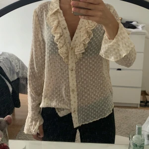 Prickig volangskjorta i beige (använd 1 gång)  - Säljer en transparent beige skjorta med prickigt mönster och volangdetaljer längs kragen och knäppningen. Skjortan har långa ärmar och knappar framtill, perfekt för dig som gillar romantiska och unika plagg.