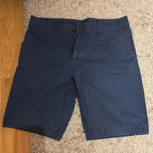 Blåa shorts från Peak Performance - Snygga marinblå shorts från Peak Performance med klassisk passform. De har fickor fram och bak, bälteshällor och stängs med dragkedja och knapp. Perfekta för sommardagar och chill häng. Materialet är mjukt och känns som bomull.