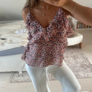 Blommig volangtop med smala axelband - Söt blommig topp med volangdetaljer och smala axelband. Toppen har en v-ringad halsringning och är perfekt för varma dagar. Färgerna går i rosa, rött och vitt med små blommor över hela tyget. Lätt och luftig känsla.