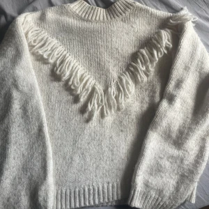 Vit stickad tröja med fransar - Mysig vit stickad tröja med långa ärmar och coola fransdetaljer i ett V-mönster över bröstet och axlarna. Perfekt för dig som vill ha en unik och trendig look med lite western-vibe. Ribbstickad i hals, ärmslut och nederkant.