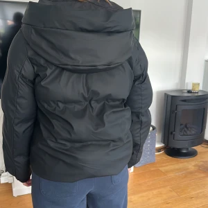 Svart pufferjacka med huva - Säljer en svart pufferjacka från veromoda med stor huva och dragkedja framtill. Jackan har två fickor med knappar men färgen på knapparna har börjat tappa färg. Perfekt för kalla dagar när du vill hålla värmen och ändå se snygg ut. Materialet påminner som en regnjacka men lite mattare🤍.