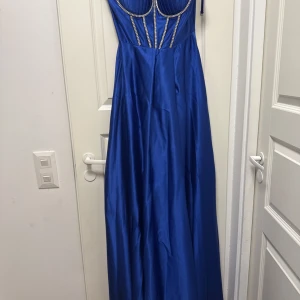 Sininen pitkä mekko strassikoristeilla - Supertyylikäs sininen pitkä mekko, jossa on kapeat olkaimet ja selkeä rintakehä. Mekossa on kimaltelevia strassikiviä vyötäröllä, jotka antavat sille tyylikkään ja elegantin ilmeen. Valmistettu kiiltävästä materiaalista, joka laskeutuu kauniisti ja tuntuu ylelliseltä. Täydellinen niille, jotka haluavat erottua joukosta!