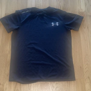 Svart Under Armour t-shirt - Svart t-shirt från Under Armour med klassisk logga på bröstet och diskret text på axeln. Modellen är enkel med rund hals och korta ärmar, perfekt för träning eller chill. Materialet är mjukt och stretchigt syntet som andas.