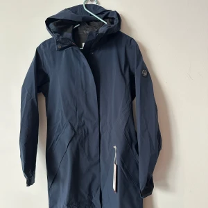 Ny Marinblå regnjacka från Sail Racing - Damjacka i polyester och spandex med tre lager. (Sail Racing Link Technology). Jackan är vattentät och vindtät med hög andningsförmåga. Handfickor med dragkedja och svetsad knappslå. Feminint skuren midja. Sail Racing-märke på ärmen och blocklogotyp upptill på huvan. 