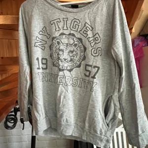 Grå sweatshirt NY Tigers Gina Tricot XS - Grå sweatshirt från Gina Tricot i storlek XS med tryck 'NY Tigers University 1957' och tigersymbol på bröstet. Långärmad modell i mjuk bomullsblandning, perfekt för chill dagar. Klassisk crewneck och avslappnad passform. Använts fåtal gånger men använder aldrig nu så hoppas den kan komma till hands för någon annan😁