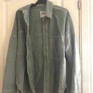 Oversized grön overshirt från Zara - Snygg oversized overshirt från Zara i en dämpad grön färg. Skjortan har klassisk krage, stora bröstfickor och knappar framtill. Tillverkad i ett kraftigare bomullstyg som ger en cool och avslappnad look. Perfekt att slänga över en t-shirt.