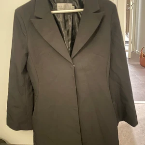 Svart balzerklänning från NLY Trend, stl 38 - Stilren svart blazerklänning från NLY Trend i storlek 38. Klassisk design med krage, knäppning framtill och slät yta. Fodrad insida i glansigt material. 