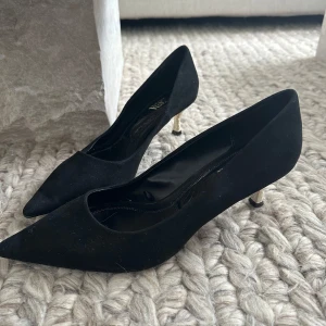 Svarta pumps med spetsig tå och guldklack - Stilrena svarta pumps med spetsig tå och låg, smal klack i guld. Skorna som är från Zara har en klassisk siluett och är tillverkade i mocka för en lyxig känsla. 