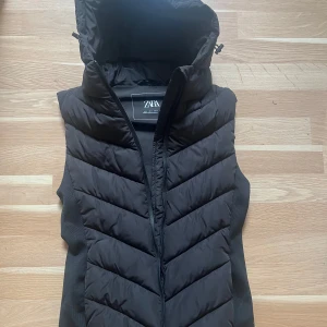 Svart dunväst från ZARA med huva - Snygg svart dunväst från ZARA med dragkedja och stor huva. Västen har ett quiltat mönster framtill och är perfekt för lager-på-lager-stil. Passar dig som gillar en sportig och clean look.