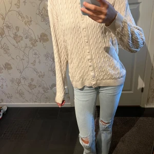 Beige kabelstickad kofta från Basic You - Kabelstickad beige kofta från Basic You i ekologisk bomull. Rund halsringning, långa ärmar och knäppning framtill med matchande knappar. Perfekt att slänga över en t-shirt eller linne för en chill och stilren look.