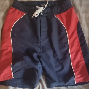 Blå och röda badshorts Black Temple - Snygga badshorts från Black Temple i marinblått med röda paneler på sidorna och vita detaljer. Shortsen har elastisk midja med snörning och innerbyxa i mesh. Perfekta för stranden eller poolen.