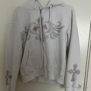 Vit hoodie med kors och strassdetaljer - Säljer en vit hoodie med dragkedja och huva. Tröjan har stora kors och vingar i strass på bröstet och mindre kors på ärmarna. Praktiska fickor framtill och snörning i huvan. Perfekt för dig som gillar statement-plagg! 