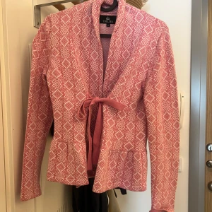 Mönstrad cardigan jacka från Chelsea - Rosa och vit cardigan jacka från Chelsea med snyggt geometriskt mönster. Jackan har knytband i midjan och långa ärmar. 