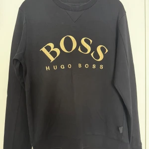 Svart sweatshirt från Hugo Boss - Svart sweatshirt från Hugo Boss med stort broderat BOSS-tryck i gult/guld på bröstet.