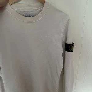 Vit långärmad tröja Stone Island - Vit långärmad tröja från Stone Island med klassisk rund hals och ikonisk logotyp-patch på vänster ärm. Tröjan har en clean look och är tillverkad i mjuk bomull, perfekt för en stilren och avslappnad outfit.