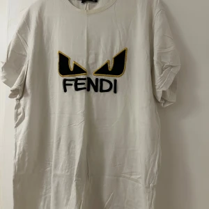 Vit Fendi t-shirt med broderad logga - Snygg vit t-shirt från Fendi med stort broderat ögonmotiv och Fendi-text på bröstet. T-shirten har klassisk passform, rund hals och är kortärmad. Materialet är mjuk bomull och detaljerna i svart och gult ger en cool kontrast.