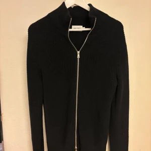 Svart stickad kofta från East West - Svart stickad kofta från East West med hel dragkedja framtill och hög krage. Tröjan har långärmad passform och är ribbstickad för en clean och stilren look. Perfekt för lager-på-lager och funkar året runt.