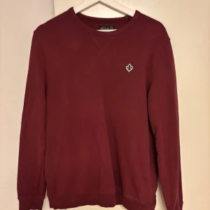 Morris vinröd sweatshirt med emblem - Vinröd sweatshirt från Morris med rund halsringning och litet broderat emblem på bröstet. Tröjan har långa ärmar och ribbade muddar vid ärmslut och nederkant. Perfekt för en chill och stilren look.