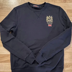 Mörkblå tröja från Morris XS - Svart sweatshirt från Morris med broderad guldfärgad logga och brittisk flagga på bröstet. Klassisk rund halsringning, ribbade muddar och lång ärm. Perfekt för dig som gillar stilren och preppy look.