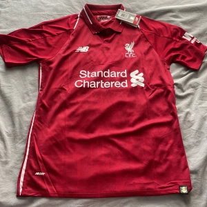 Liverpool FC röd matchtröja New Balance - Liverpool FCs officiella hemmatröja från New Balance i rött med vita detaljer. Klassisk pikékrage, korta ärmar och broderat klubbmärke på bröstet. Tillverkad i lätt och ventilerande polyester med tryckt sponsorlogga framtill.